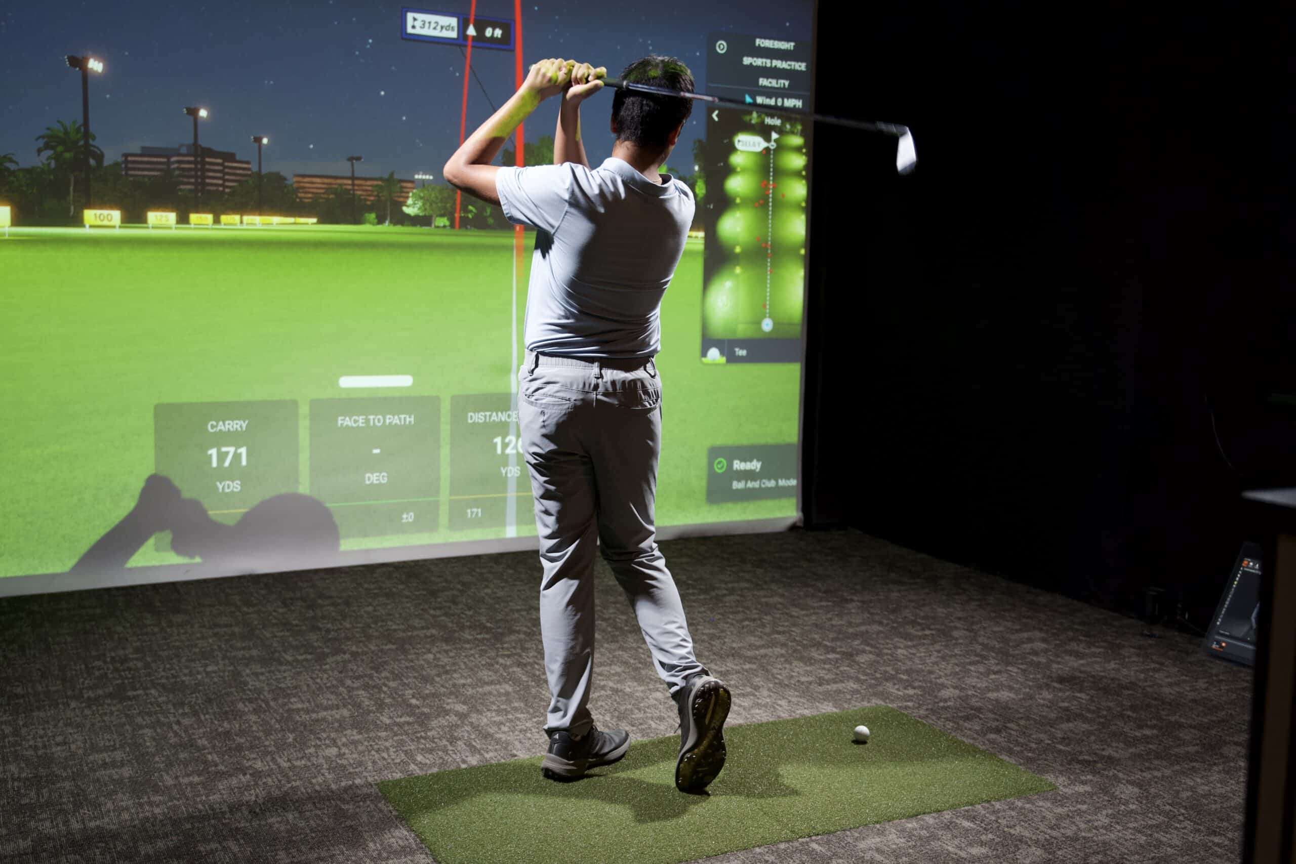 Person swings a golf club in a simulated indoor driving range with a large projected screen showing a golf course.